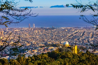 Fotografía en color de la ciudad de Barcelona. Se ha tomado desde un alto y se ve la ciudad en primer plano y al fondo, el mar.